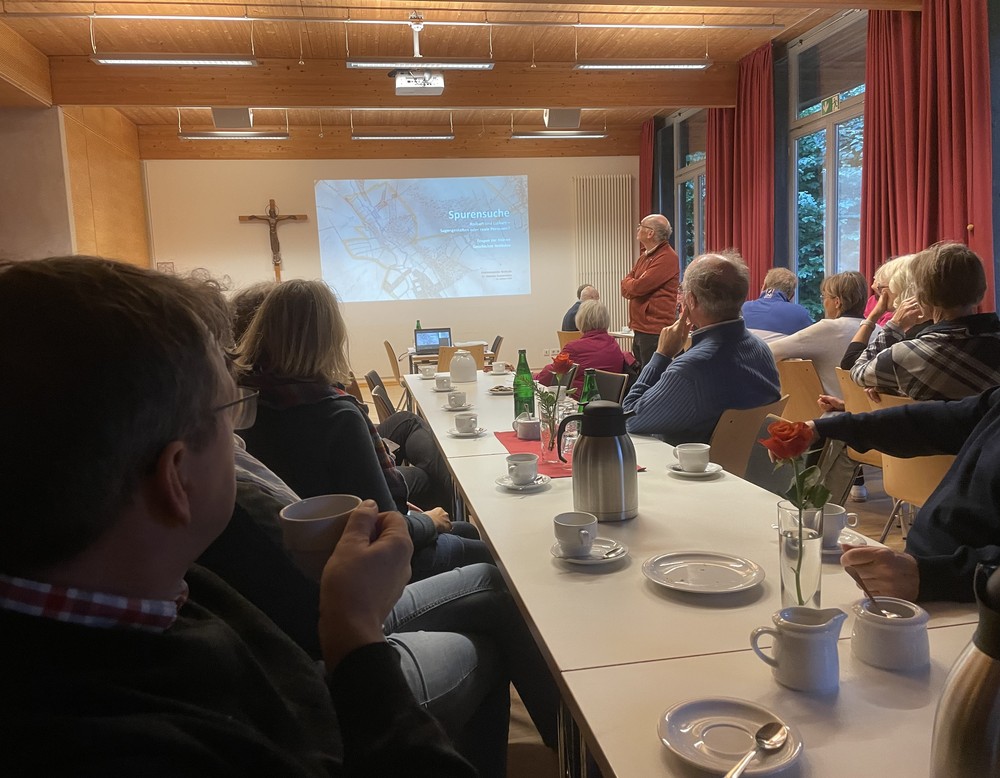 Dr. Mathias Austermann hält einen Vortrag mit PowerPoint-Präsentation. Menschen sitzen im Vordergrund an Tischen mit Kaffe und Kuchen.